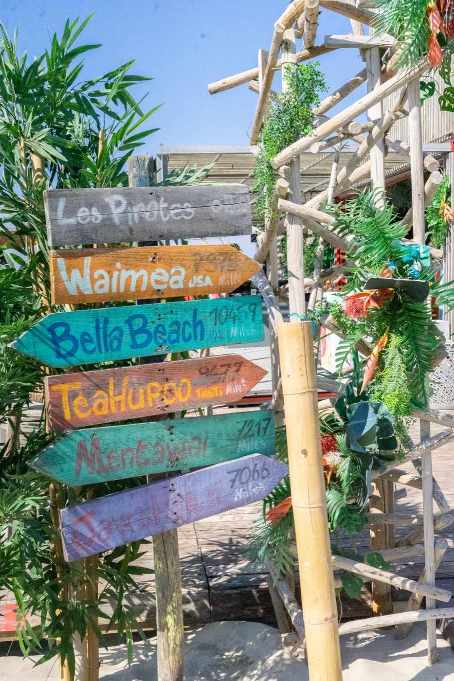 Le poteau indicateur en bois peint de La Plage des Pirates : « Les Pirates », Waimea, Bella Beach, Teahupoo, Mentawai, entre palmes et bambous, sous un ciel bleu.